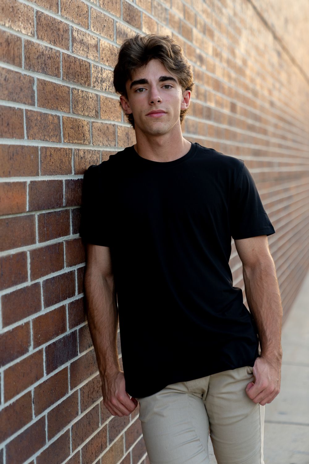 Young man in black t-shirt leaning against a brick wall in a casual outdoor portrait.