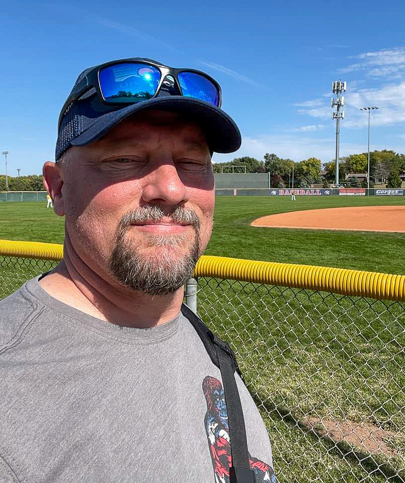 Man wearing sunglasses and cap taking a selfie at a baseball field on a sunny day.
