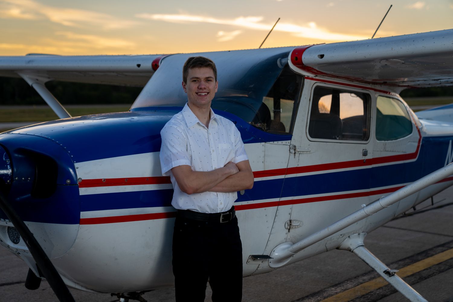 Young male pilot standing with arms crossed in front of a classic blue and white Cessna airplane at sunset.