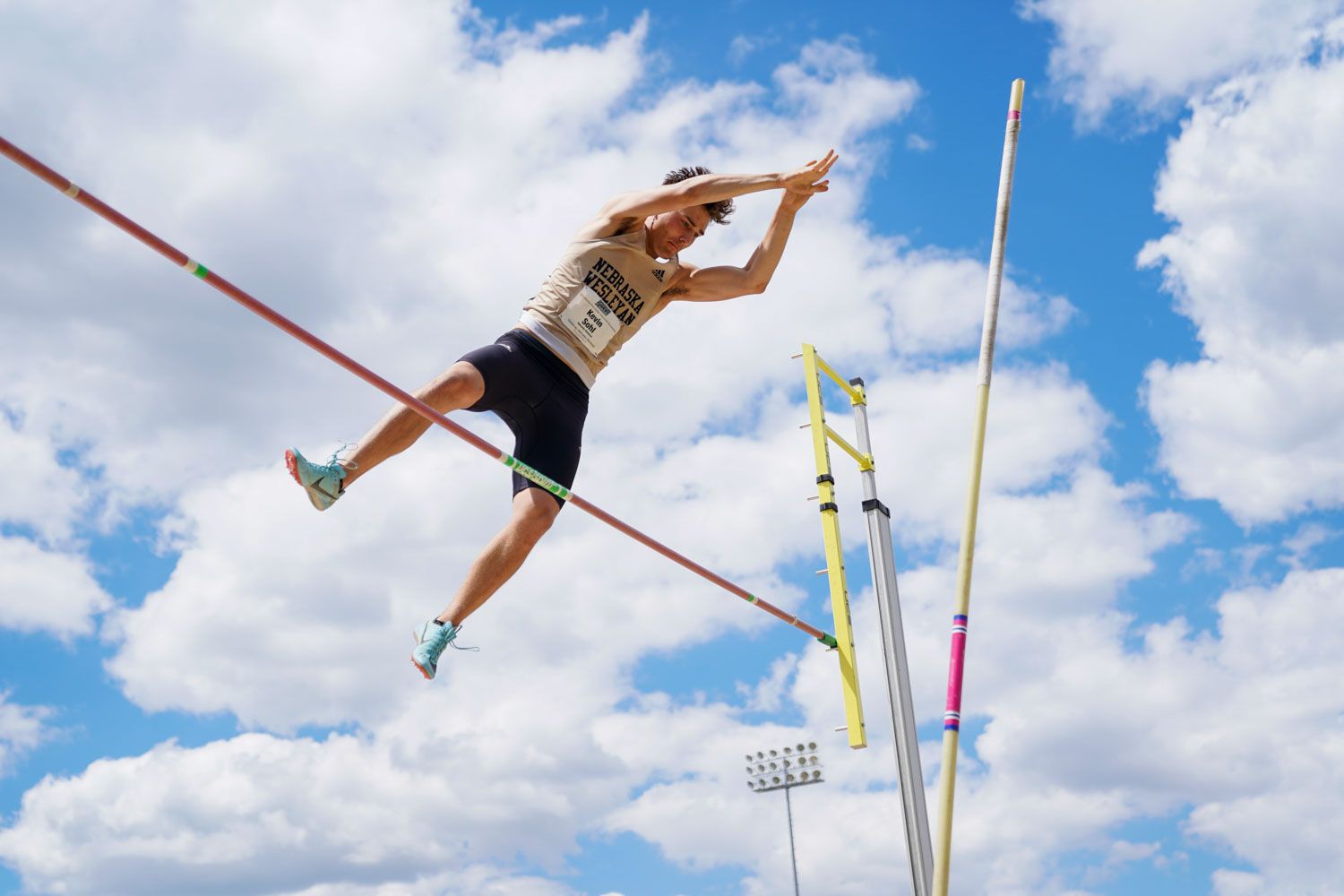 Male athlete clears pole vault bar against bright blue cloudy sky during track meet.