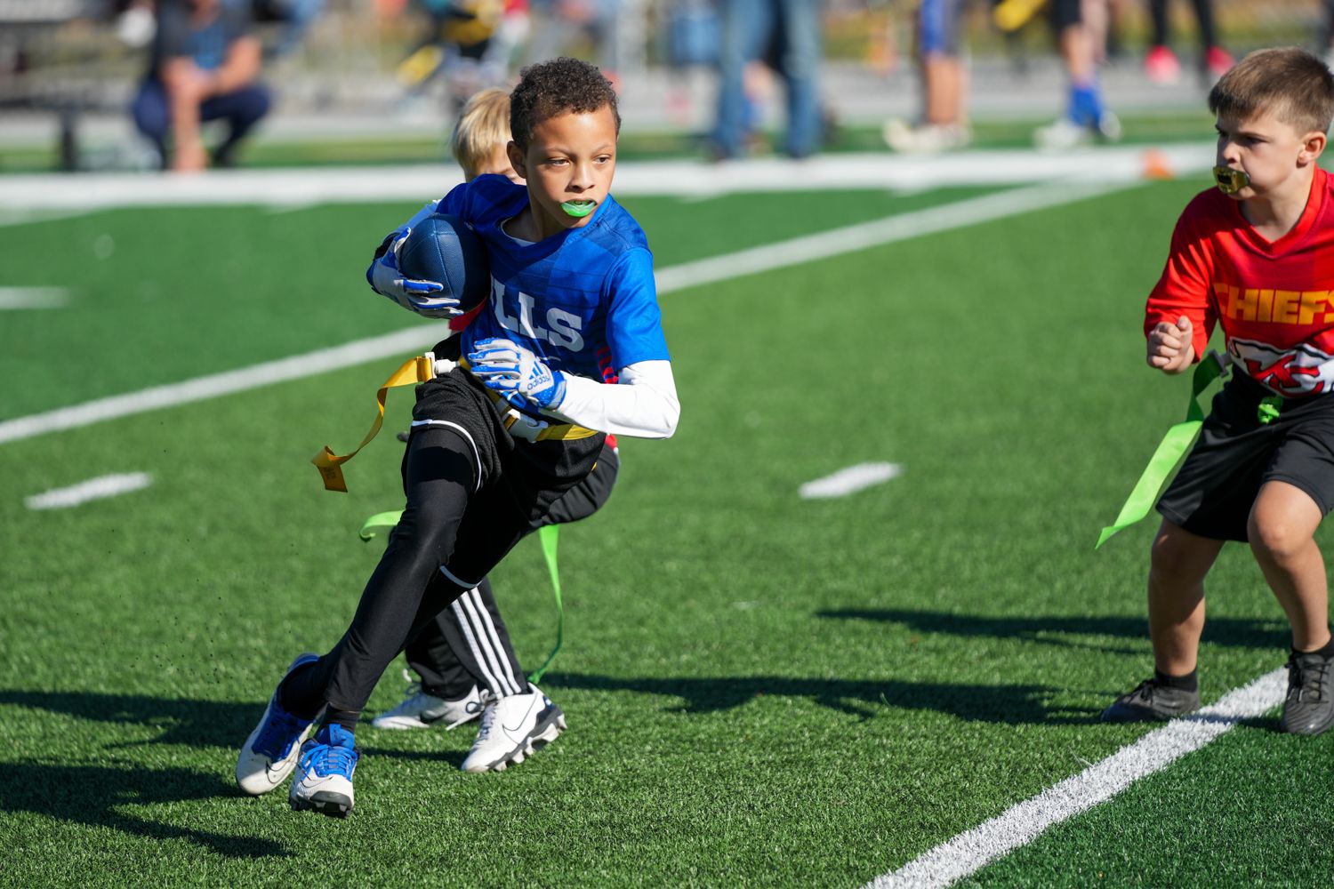 Young boy in blue jersey sprinting with football during youth flag football game.