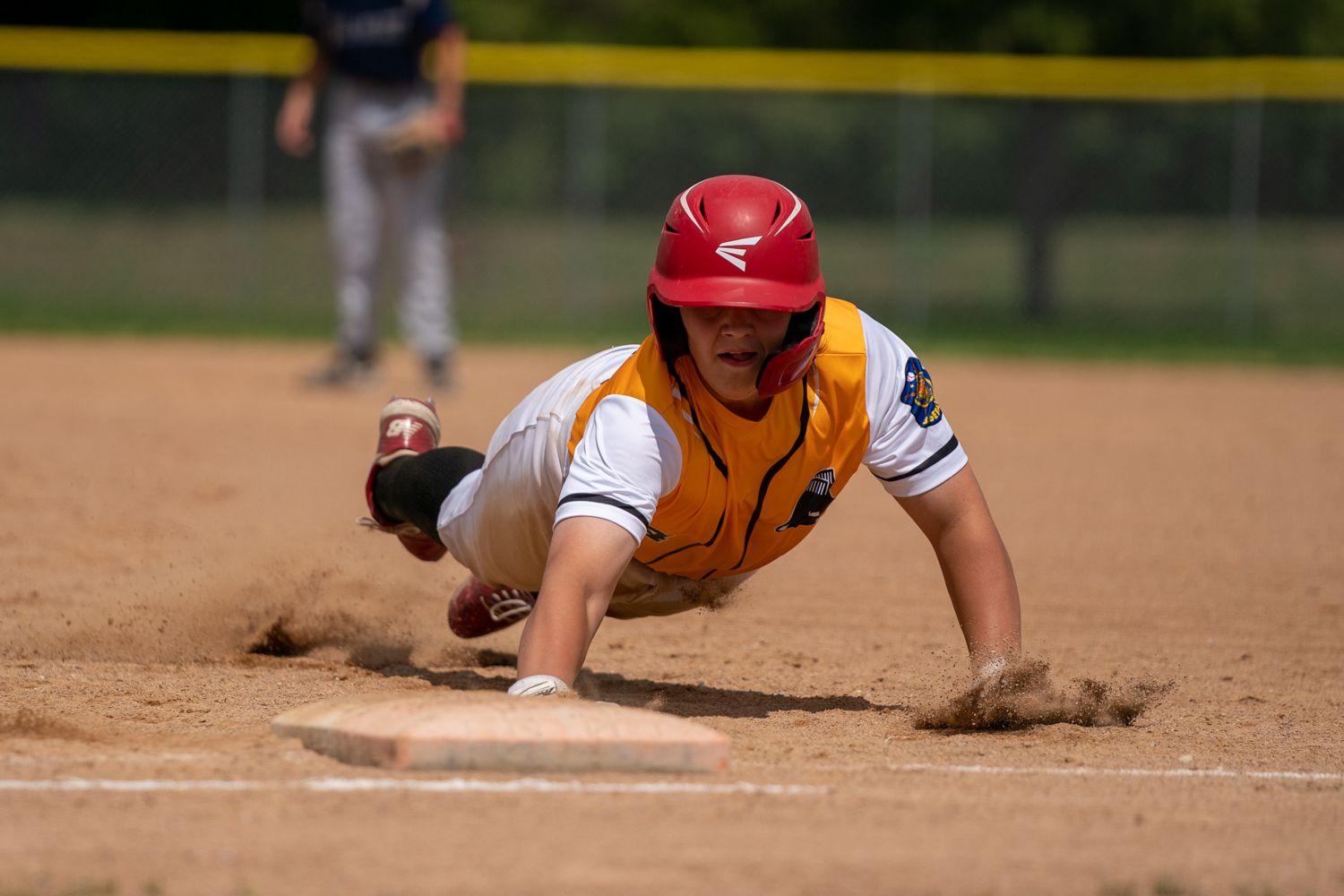 Baseball player in red helmet dives headfirst into base during game on dirt field.