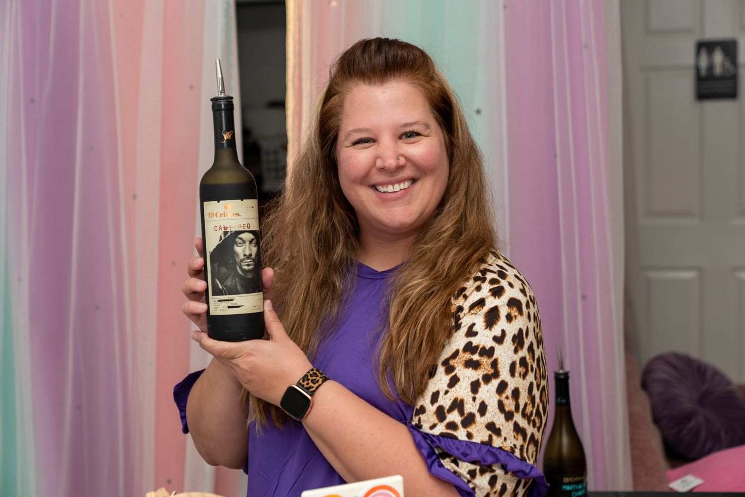 Smiling woman in leopard print cardigan proudly holds a bottle of red wine at an indoor social event.
