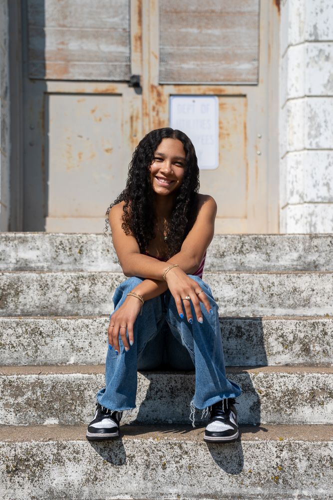 Young woman with curly hair smiling, sitting on stone steps in jeans and sneakers outdoors.