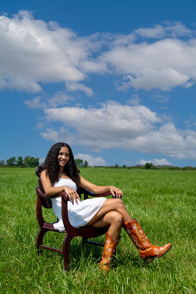 Young woman in white dress and cowboy boots sitting in wooden chair in green field under blue sky.