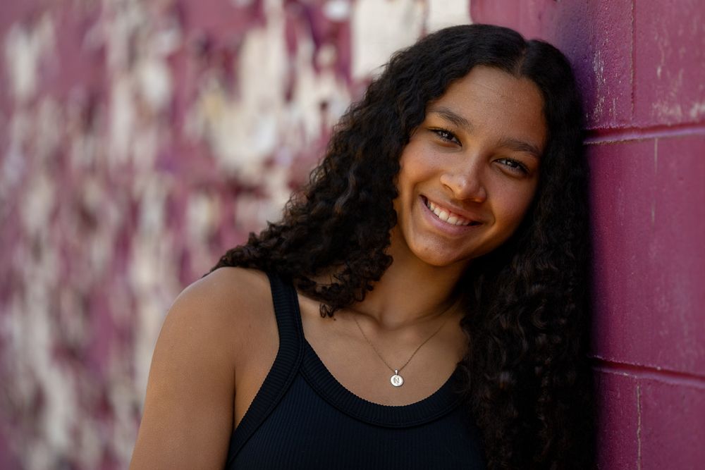 Young woman with curly hair smiling against a rustic purple wall, wearing a black tank top and delicate necklace.