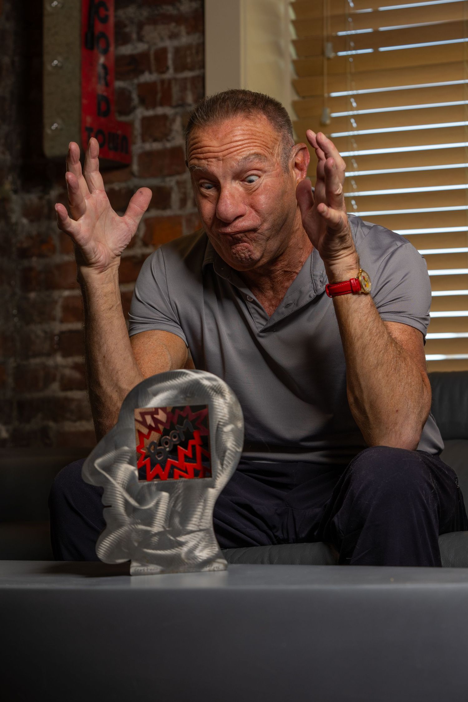 Expressive older man in gray shirt gesturing with hands raised beside a trophy.