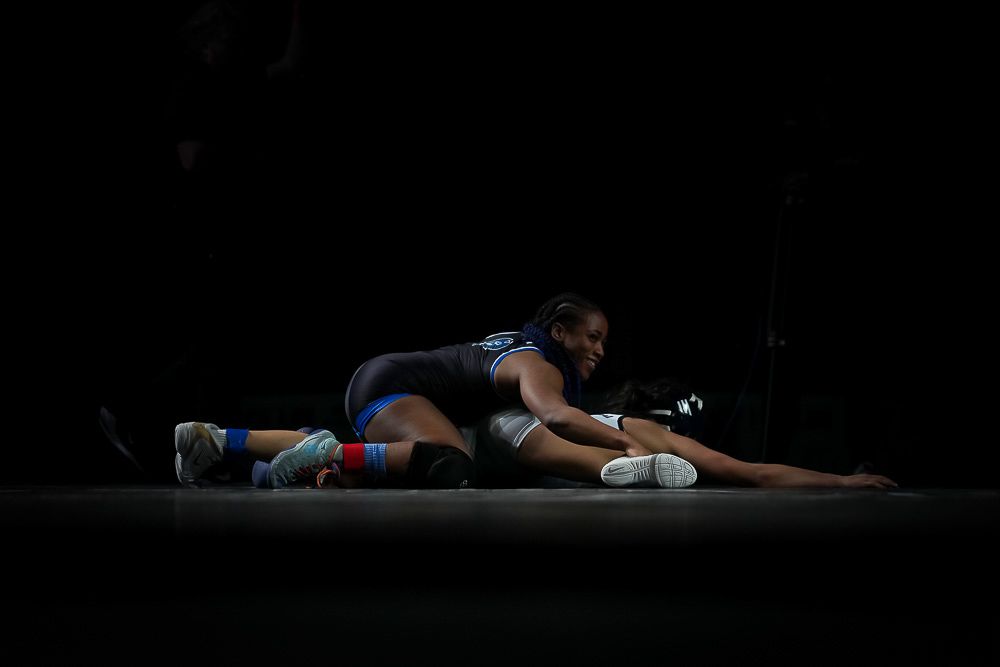Two wrestlers compete on a mat in a dimly lit arena, one pinning the other to the ground.