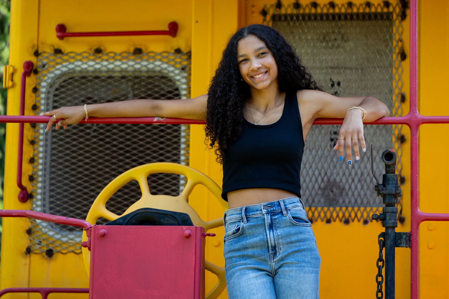 Smiling young woman in black crop top and jeans posing against colorful yellow and red playground equipment.