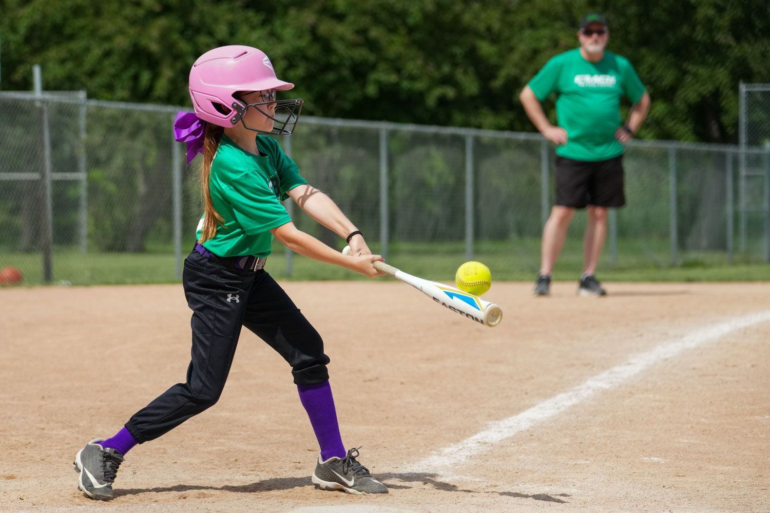 Young girl in green uniform swings at yellow softball during youth league game.