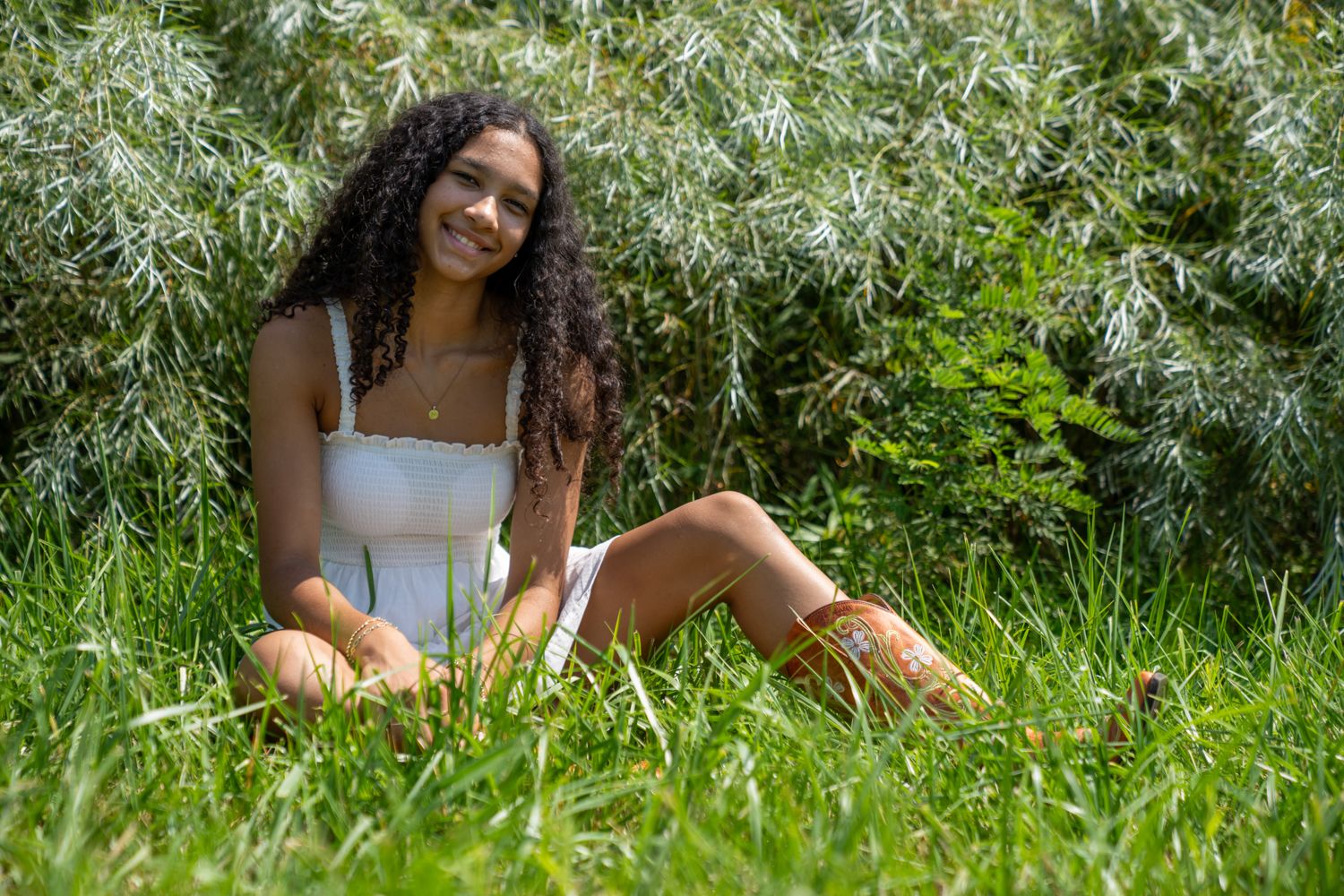 Smiling young woman with curly hair in white dress sitting in lush green grass near leafy shrubs outdoors.