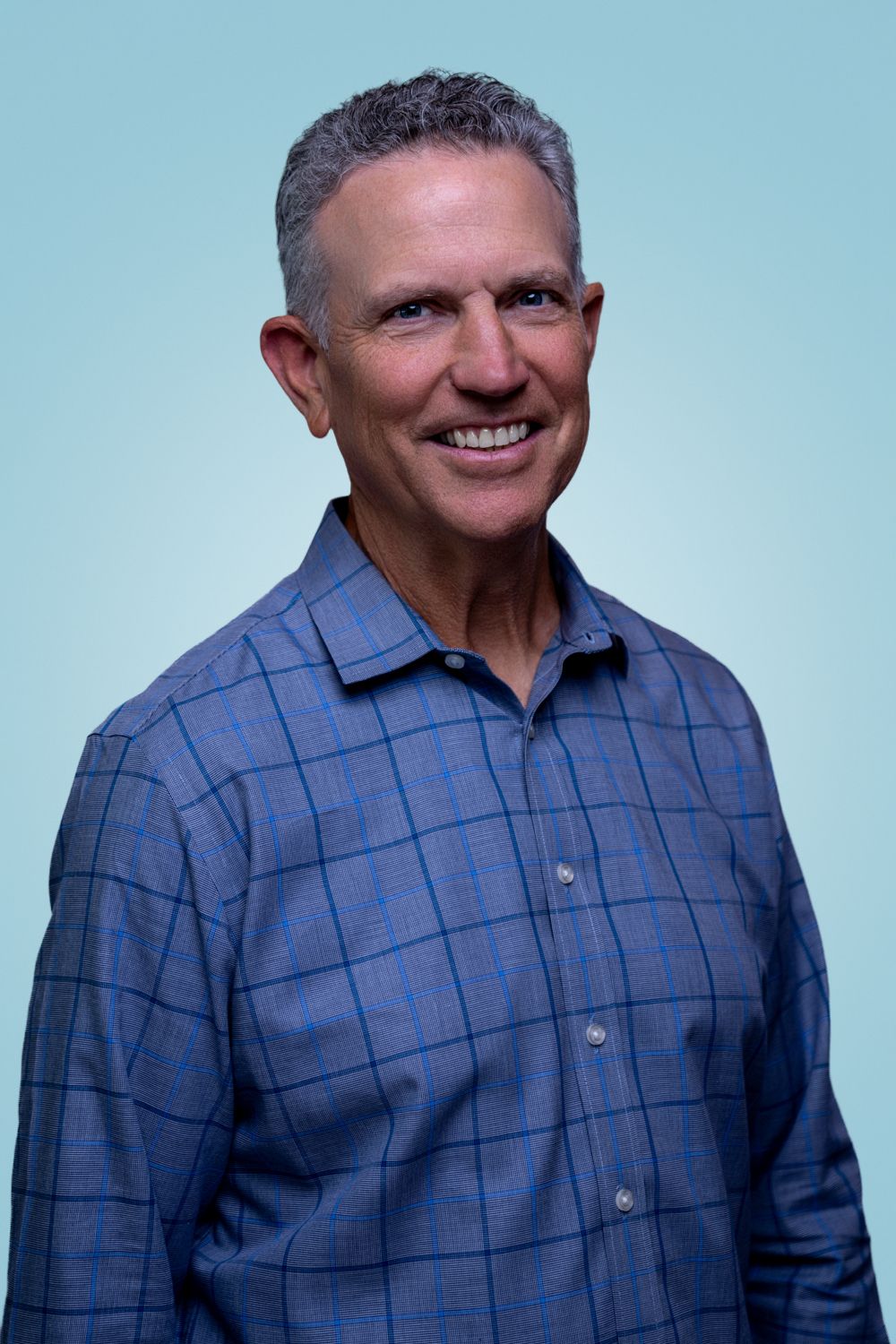 Fit middle-aged man with gray hair in blue plaid shirt smiling against light blue background.
