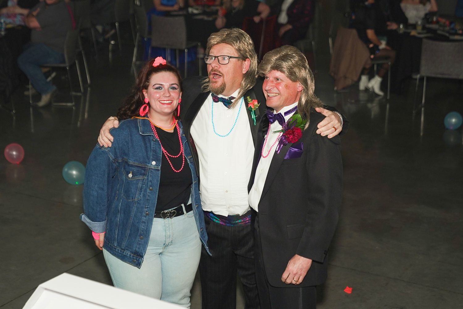 Three people in colorful retro costumes posing together at a themed party with balloons in background.