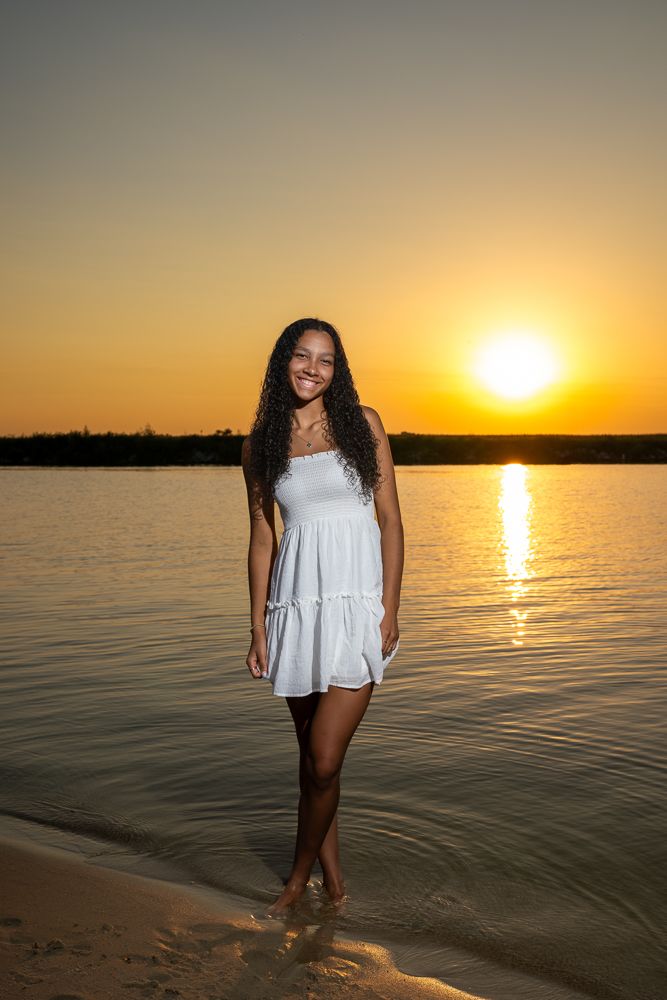 Young woman in white sundress smiling barefoot on sandy beach shore at golden sunset over calm water.