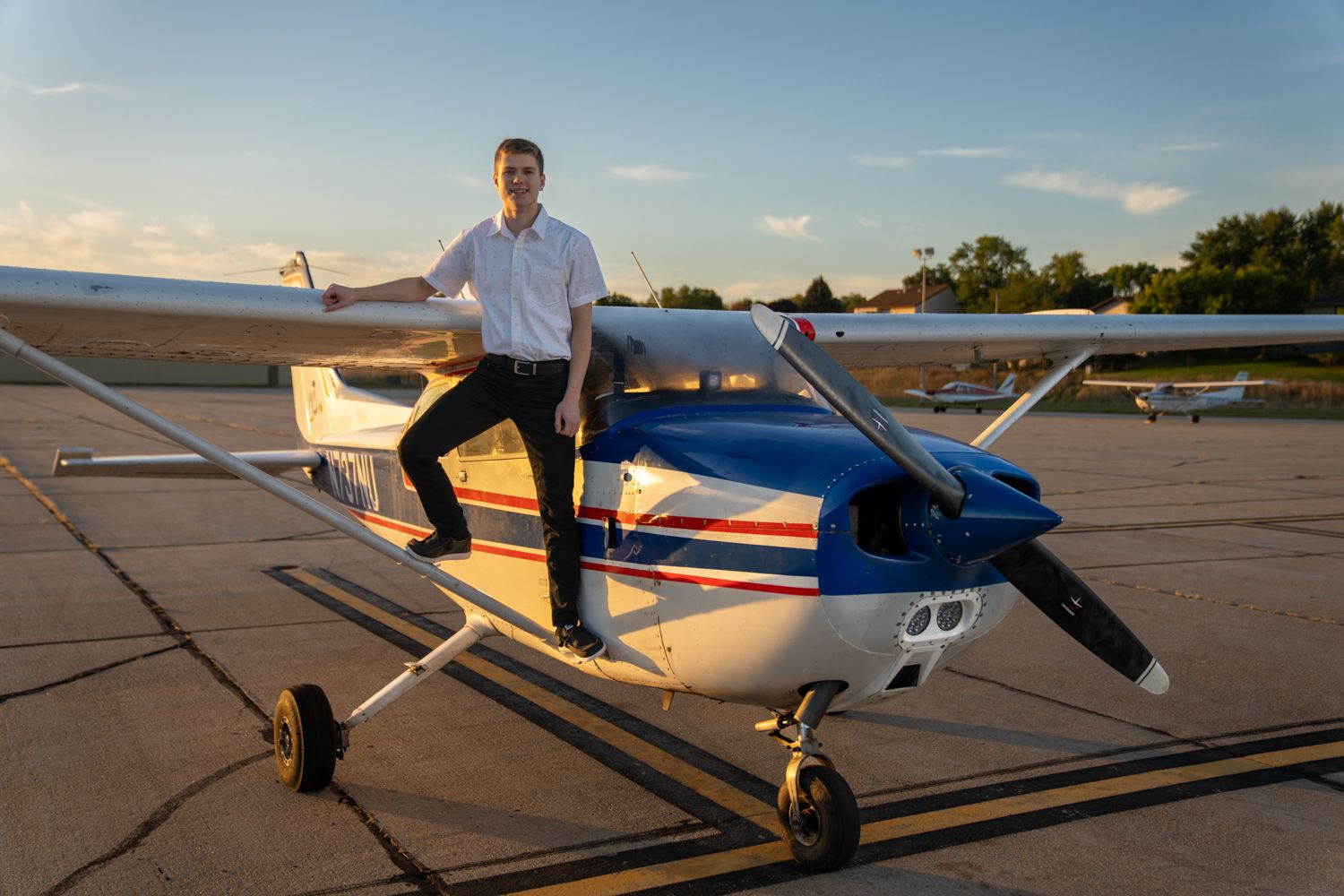 Young man in white shirt posing confidently beside a blue and white small aircraft on a runway at sunrise.