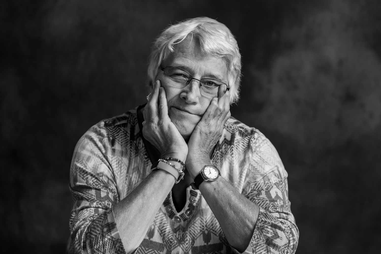 Black and white portrait of an elderly woman with glasses resting her face in her hands, thoughtful expression.
