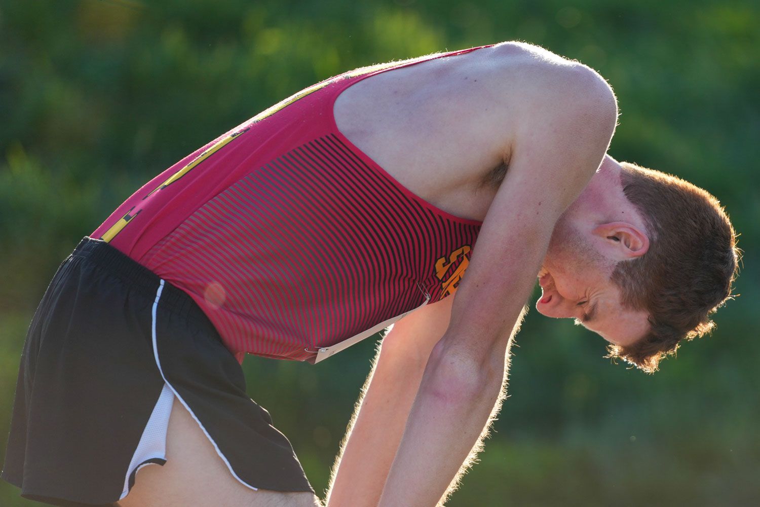 Male track and field athlete in a red uniform bending forward in exhaustion or stretching after a race outdoors.