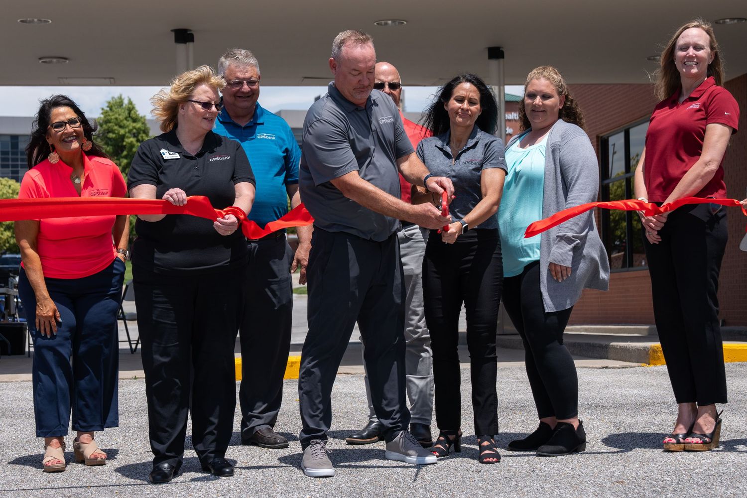 Group of people cutting a red ribbon at a grand opening ceremony outdoors on a sunny day.