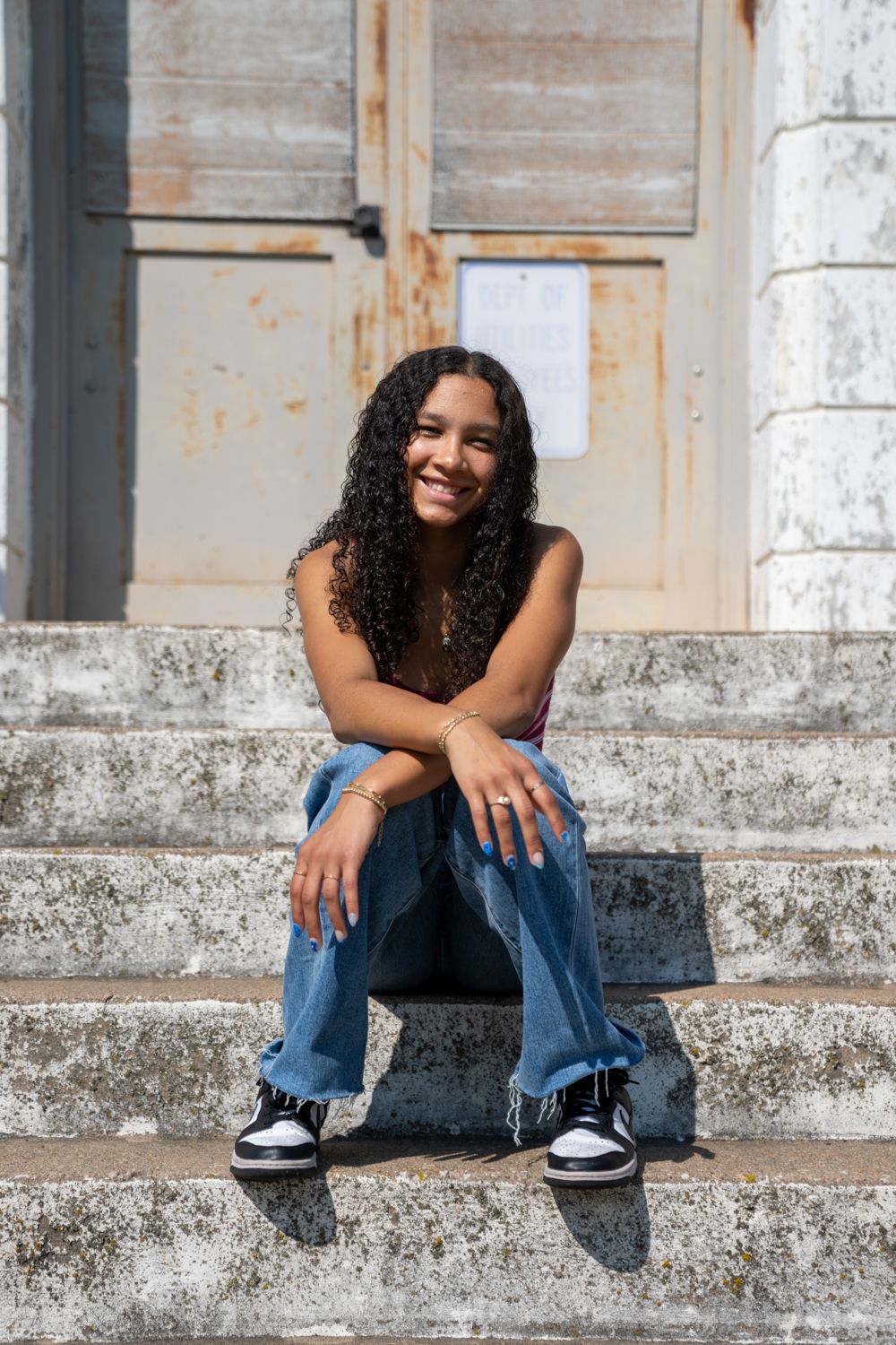 Smiling young woman with curly hair in jeans sitting on concrete steps of old building.