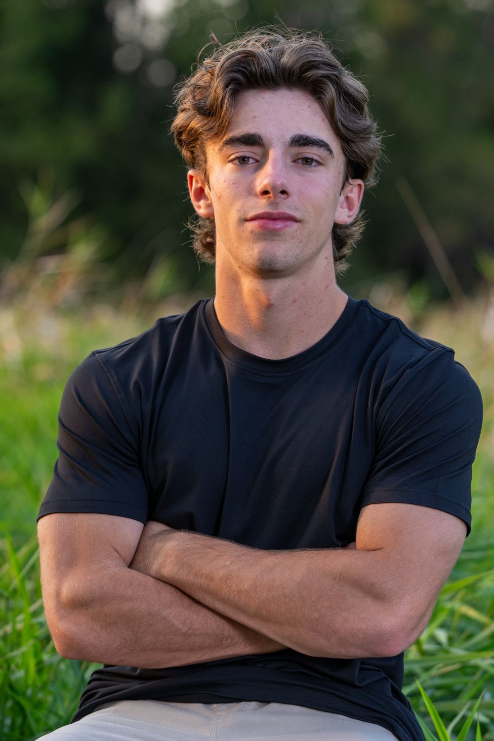 Confident young man in black t-shirt sitting with arms crossed in a natural green outdoor setting.