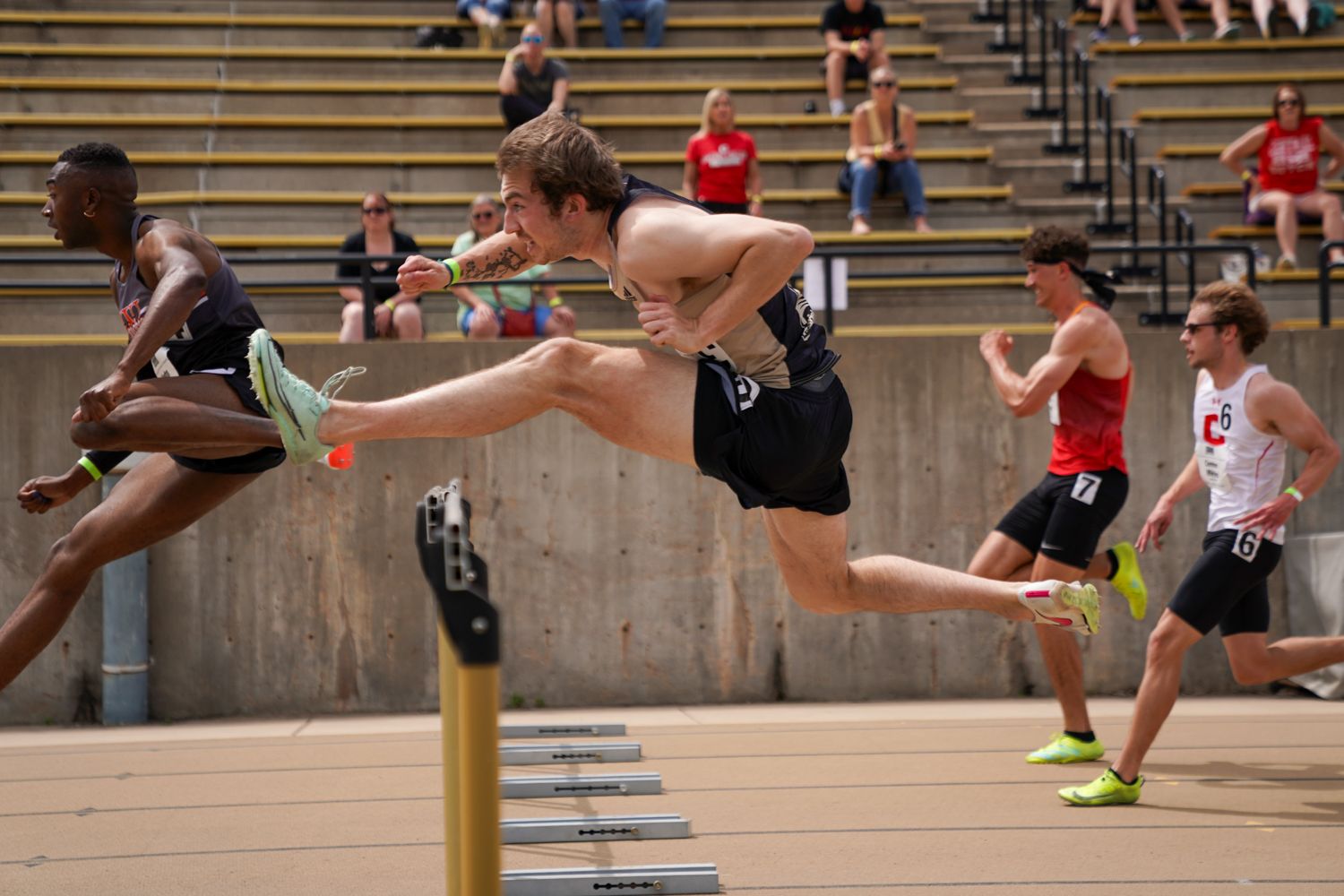 Male athletes competing in a hurdles race at an outdoor track and field event in a large stadium.