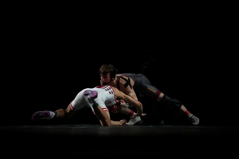 Two wrestlers compete intensely on a dark mat, one in white and one in black uniform, in a dramatic low-light setting.