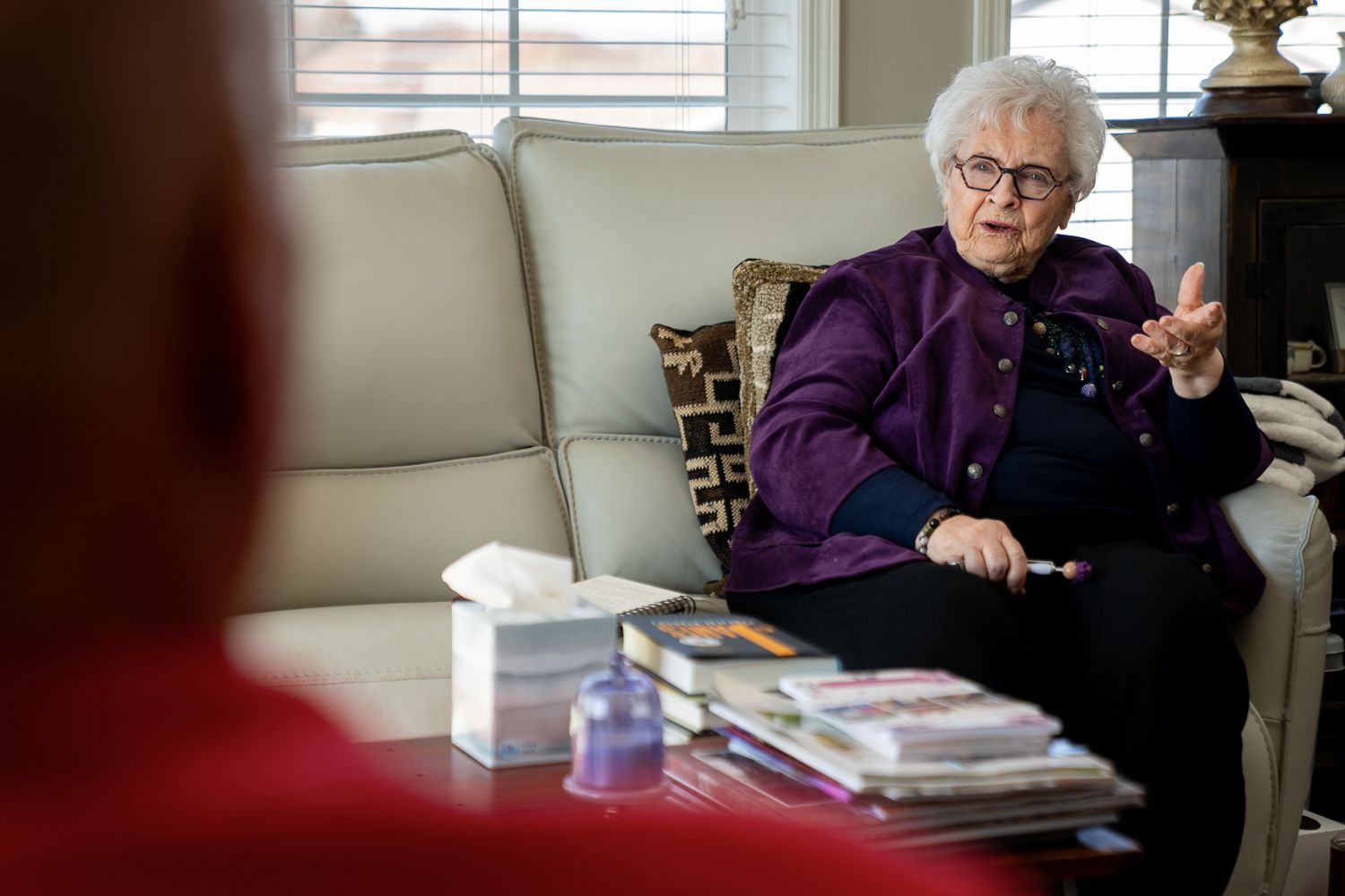 Elderly woman in purple jacket gesturing while speaking seated on white sofa indoors.