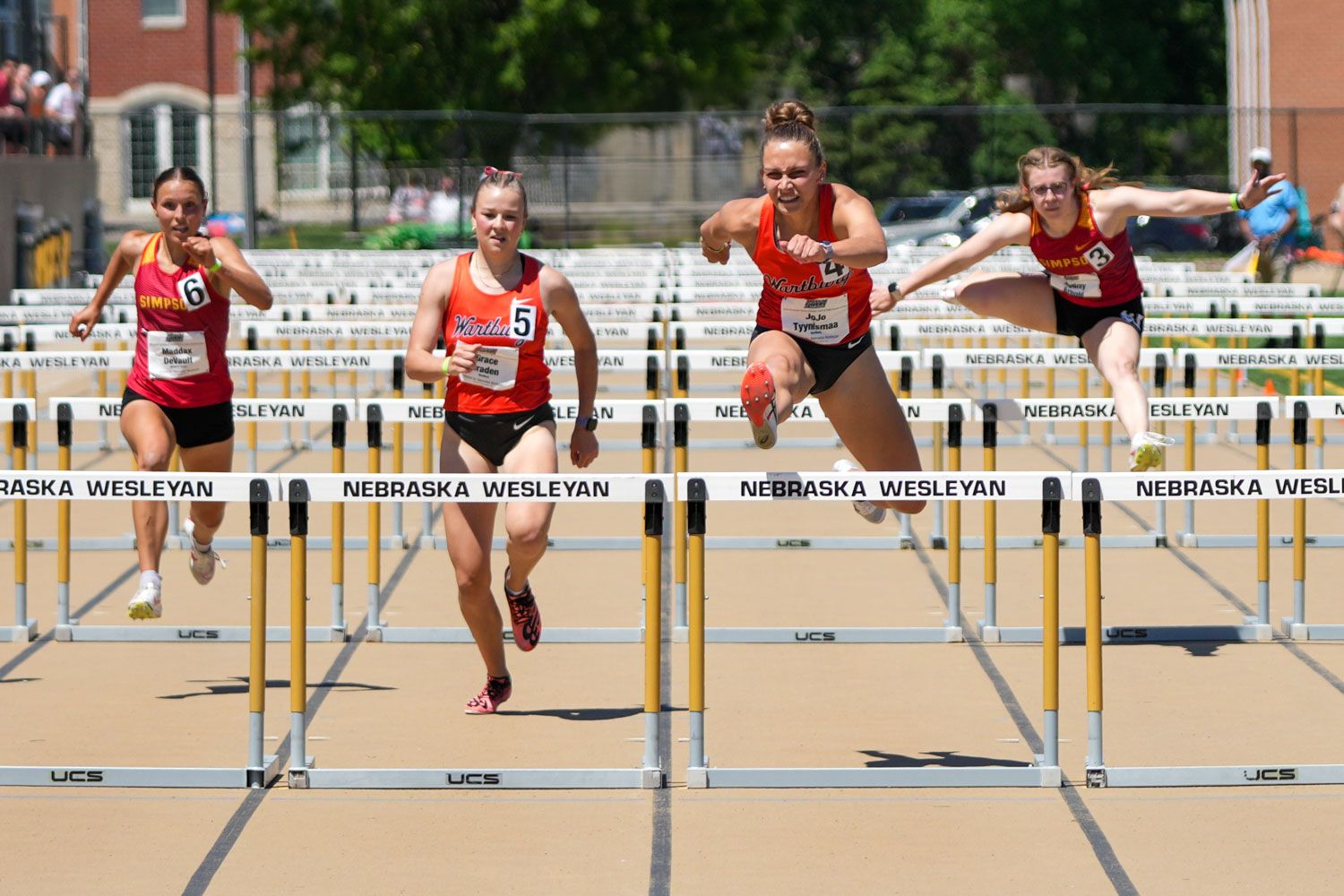 Four female athletes competing in a hurdles race at a Nebraska Wesleyan outdoor track and field meet.