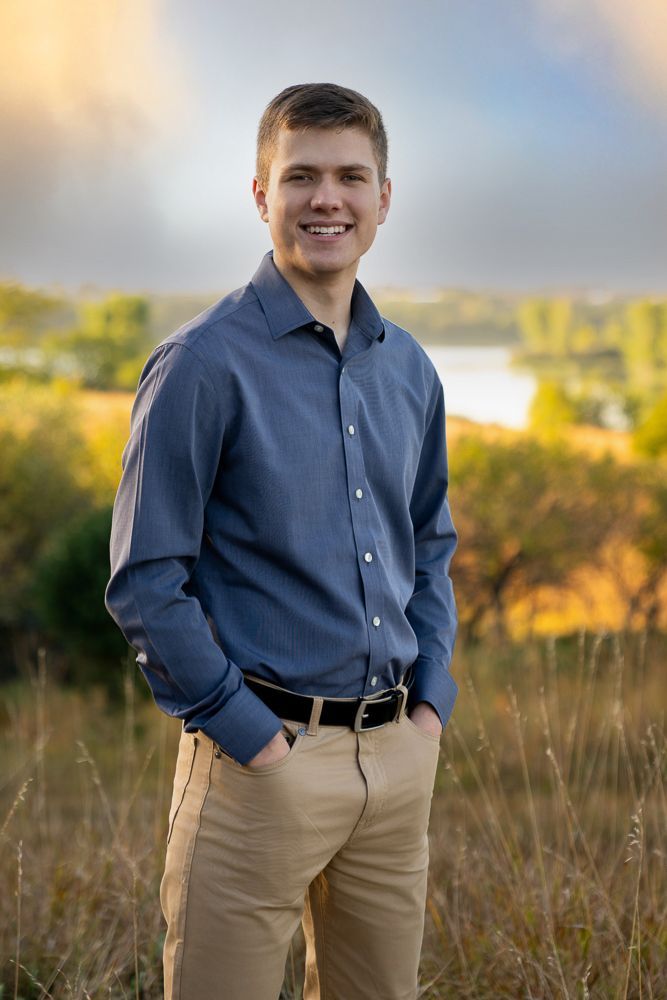 Smiling young man in blue shirt and khaki pants standing in a field with a lake and colorful sky backdrop.