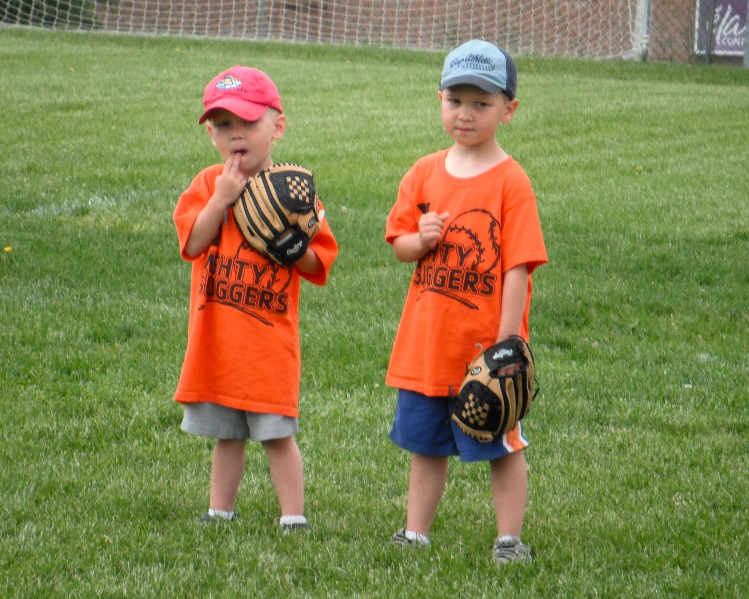 Two young children in orange shirts holding baseball gloves on green field.