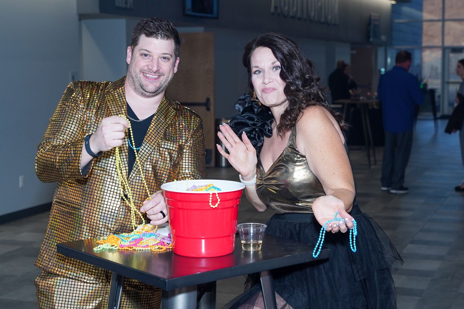 Man in gold sequin jacket and woman in black dress standing at a table with a red bucket at an event.