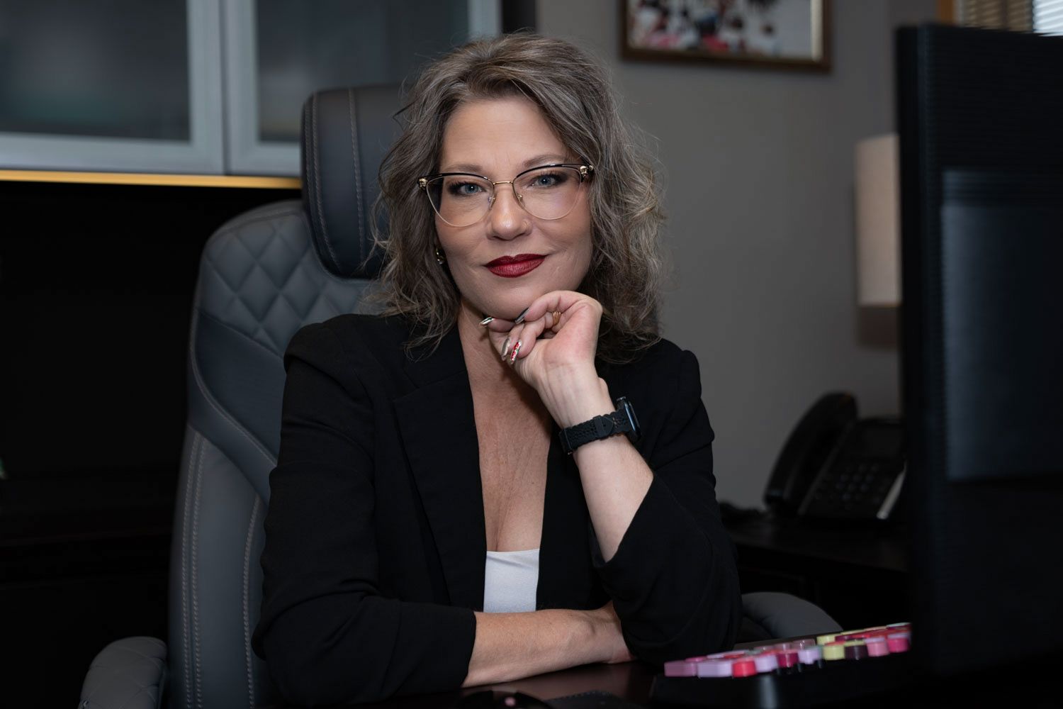 Professional woman in black blazer sitting at desk with hand on chin in office setting.