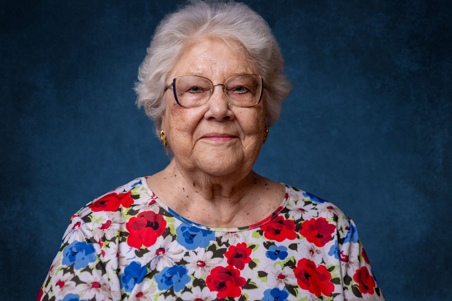 Senior woman with white hair and glasses wearing a colorful floral blouse smiling warmly.