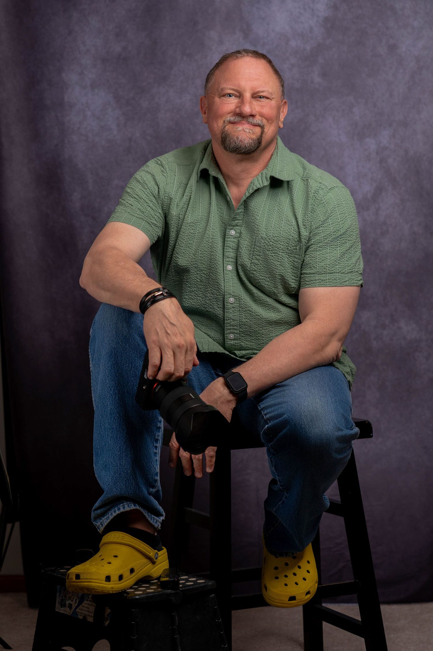 Smiling man in green shirt and yellow Crocs sitting on a stool in a photography studio.