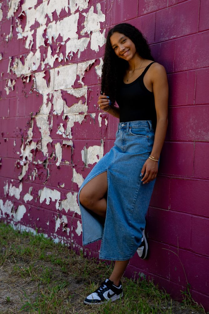 Young woman in black tank top and denim midi skirt smiles leaning against a peeling painted brick wall.