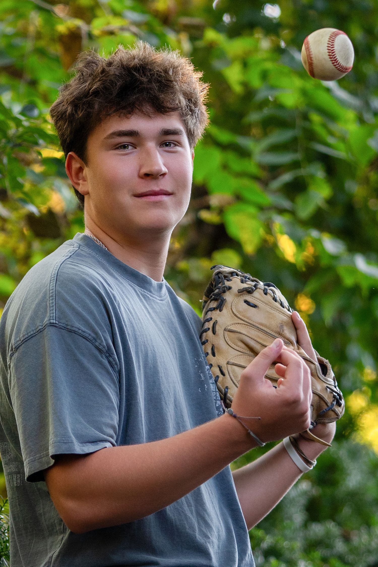 Teen boy with baseball glove tossing a baseball in the air outdoors among green trees.