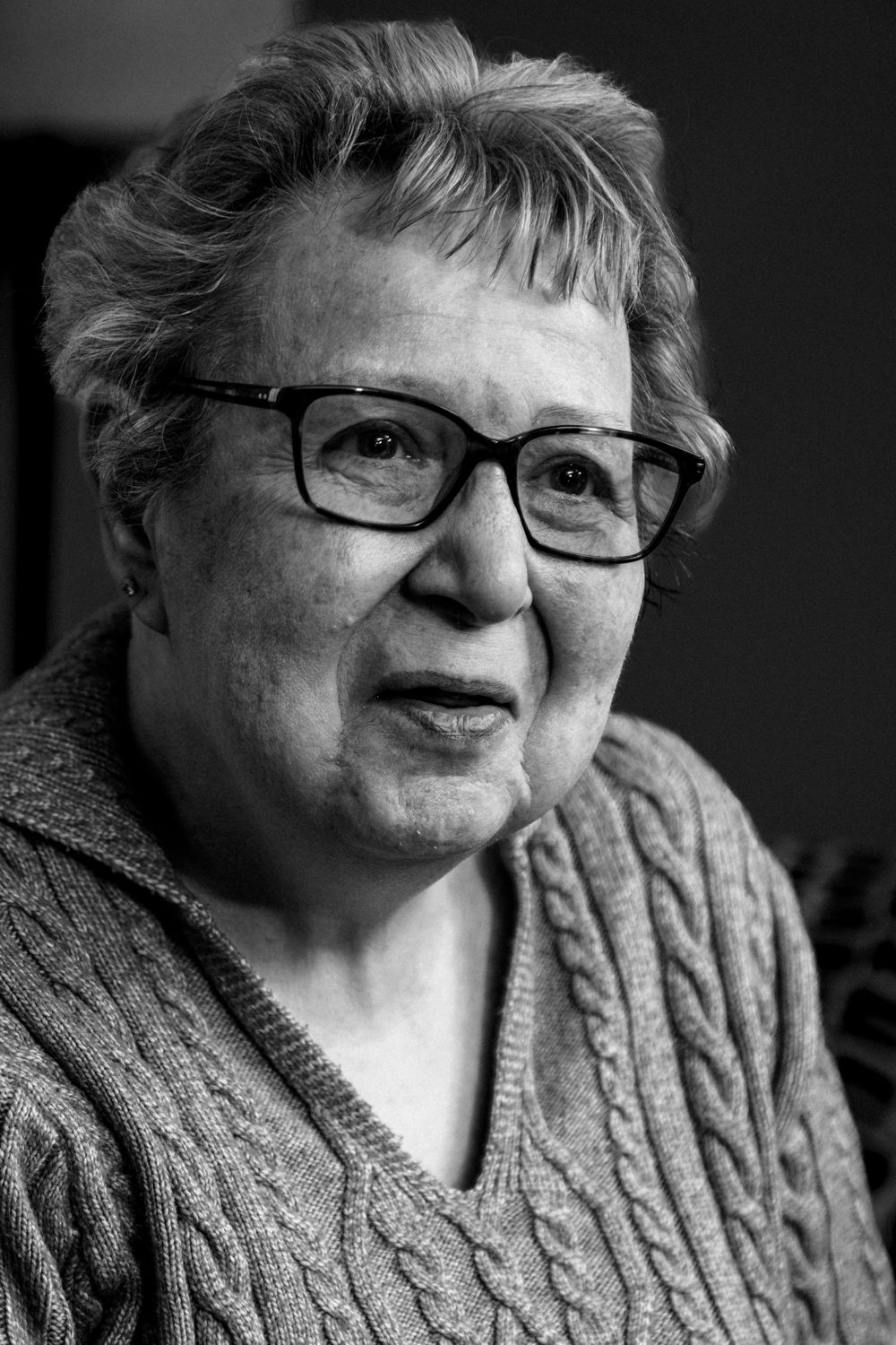 Black and white close-up portrait of an elderly woman with glasses in a cable knit sweater.