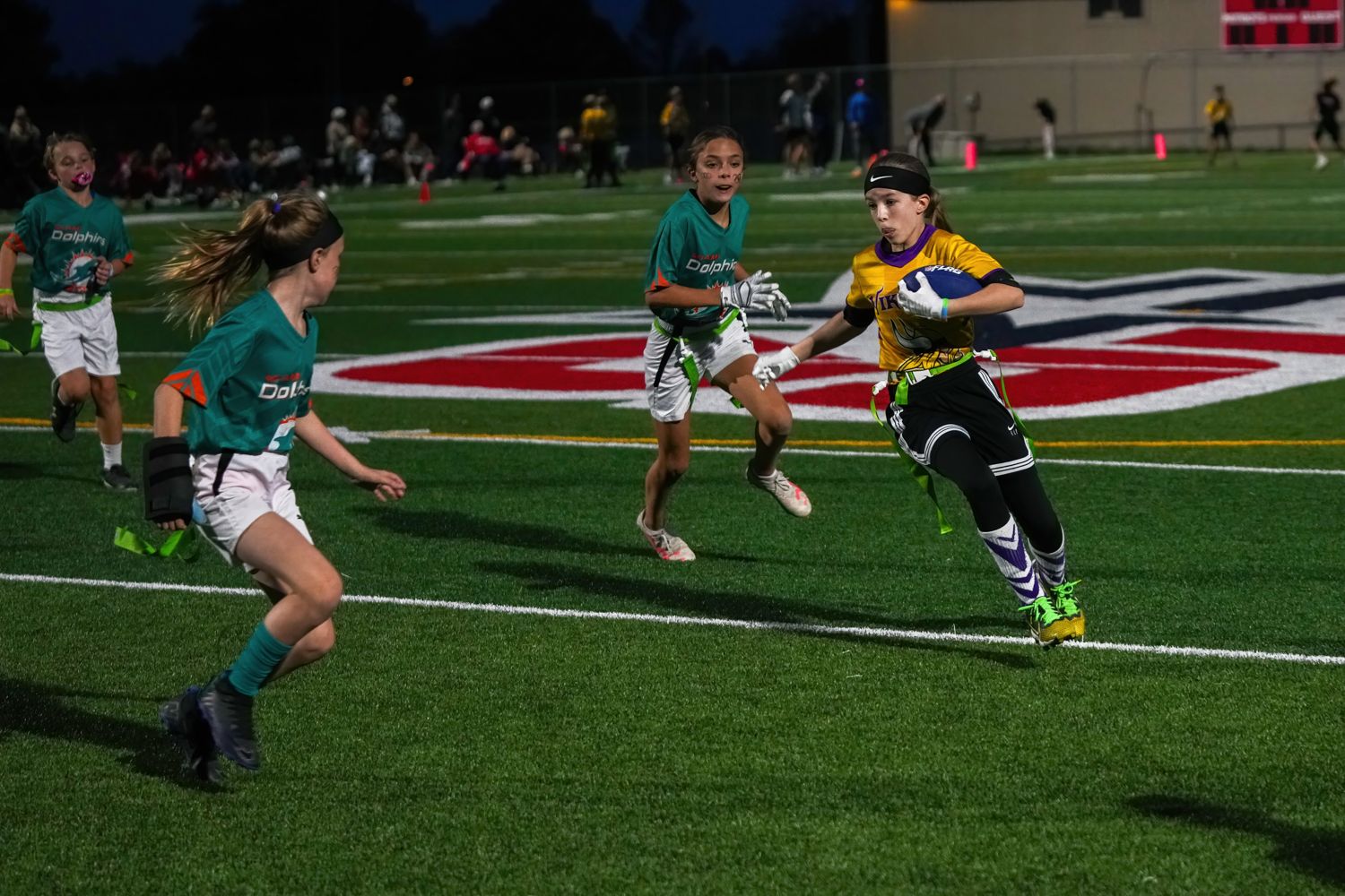 Female flag football player in yellow jersey running with ball on turf field at night.
