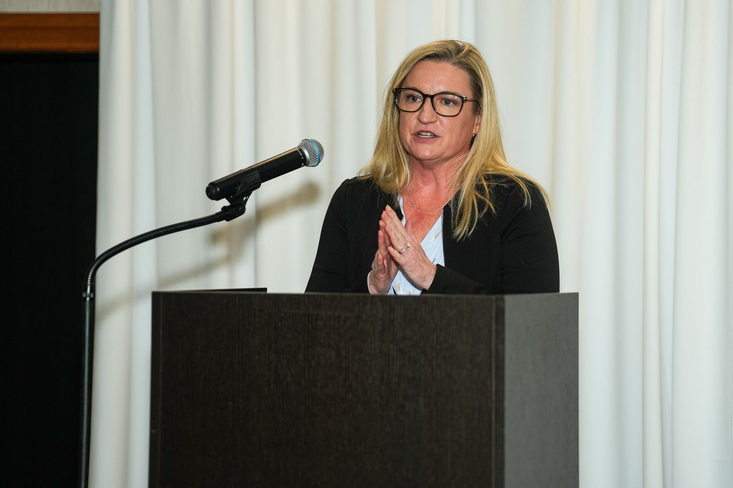 Woman with blonde hair speaking at a dark wooden podium with a microphone at a formal event.