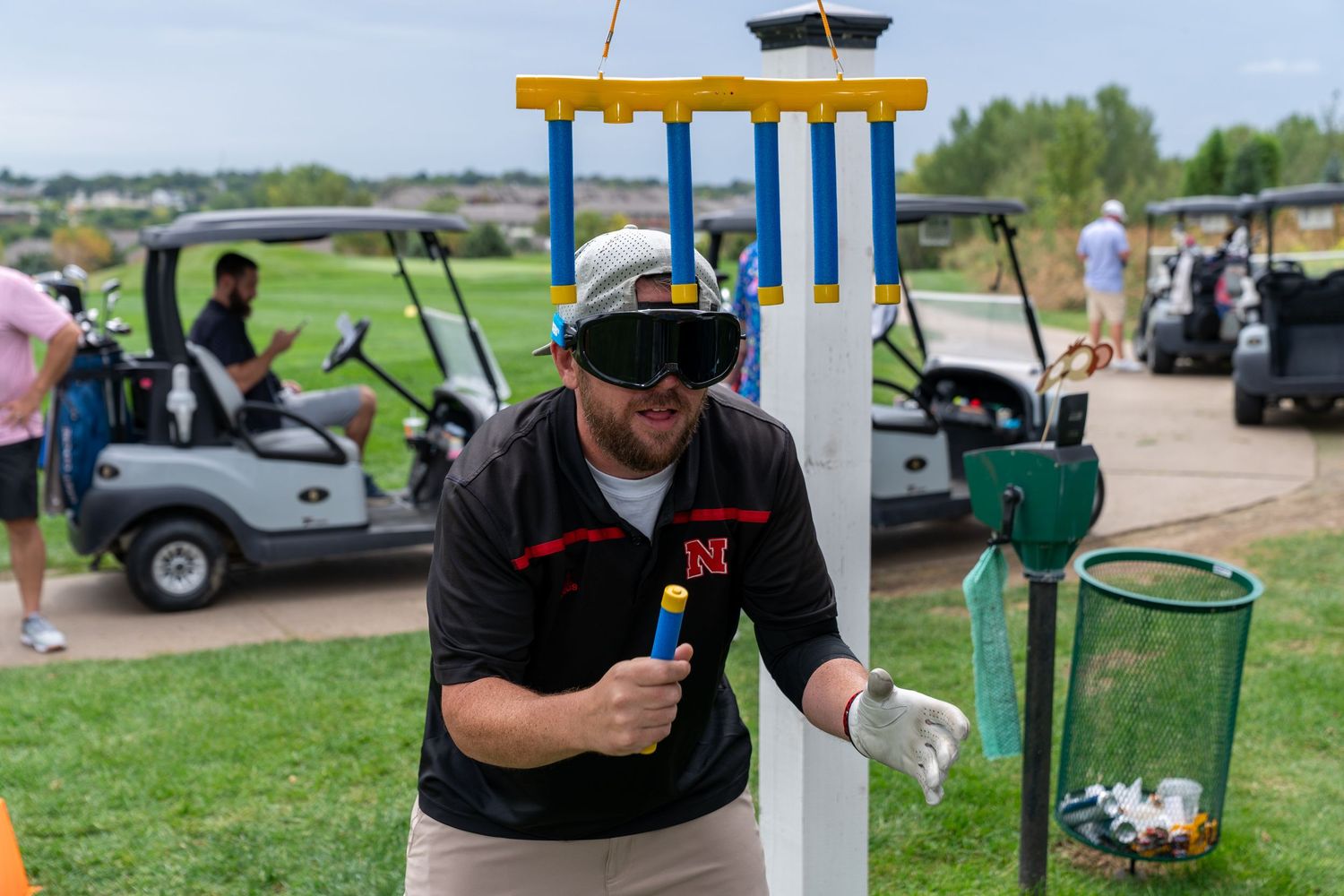 Man in baseball cap and blindfold trying to catch falling tubes at outdoor golf charity event with golf carts in background.