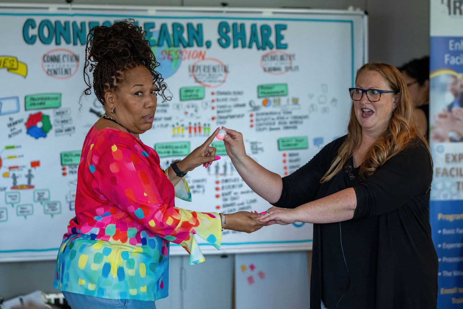 Two women exchange items in front of a Connect Learn Share whiteboard presentation.