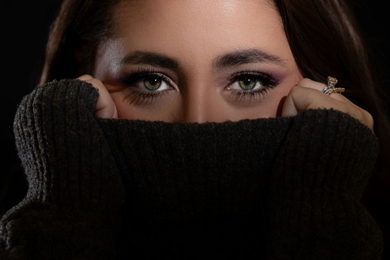 Close-up of woman with green eyes hiding face behind black turtleneck sweater.