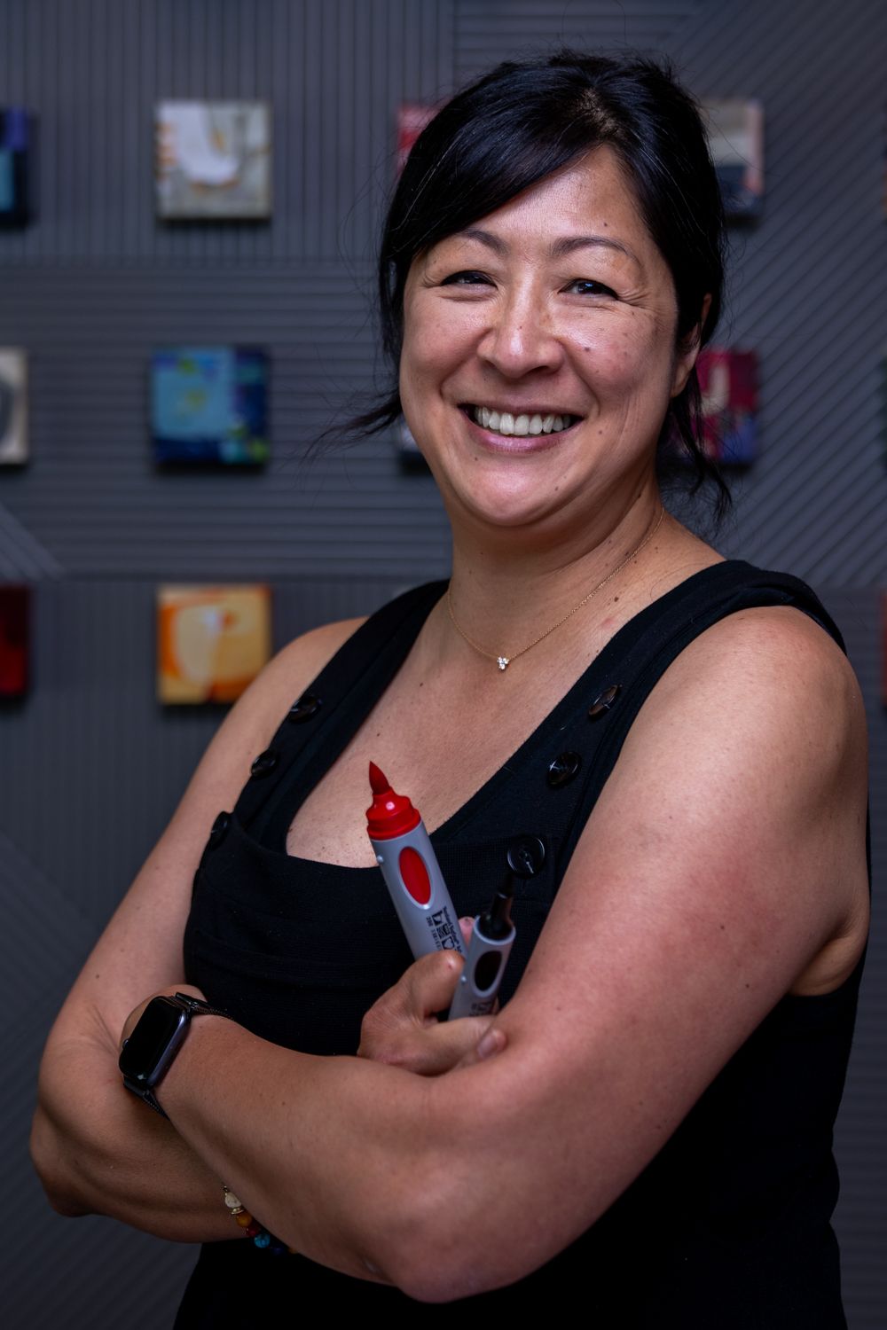 Smiling Asian woman in black sleeveless top holding a marker with arms crossed in front of colorful artwork.