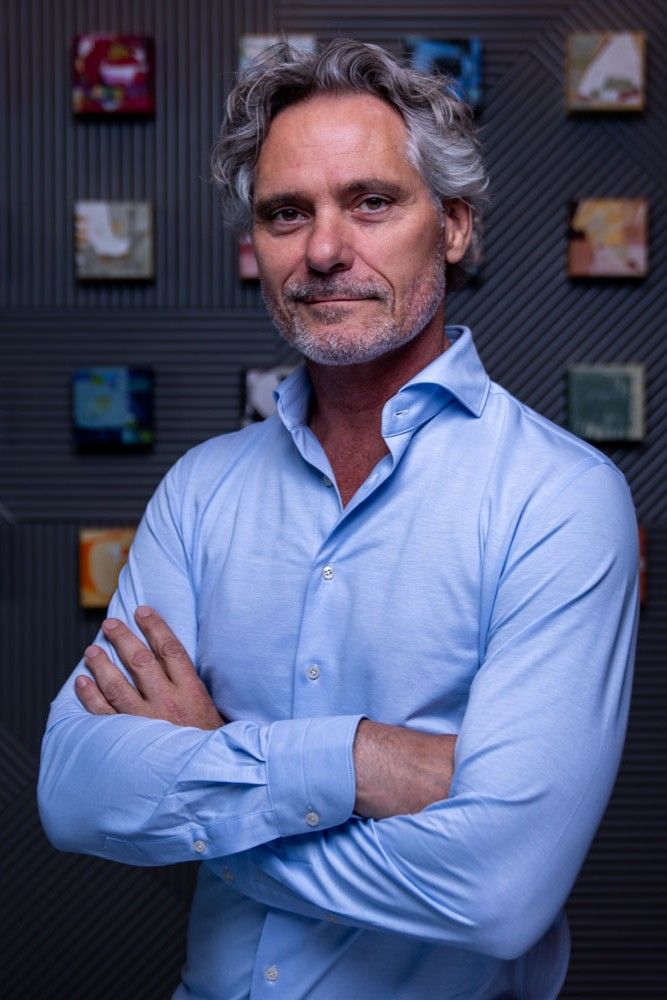 Middle-aged man with gray hair and beard in blue shirt stands with arms crossed confidently.