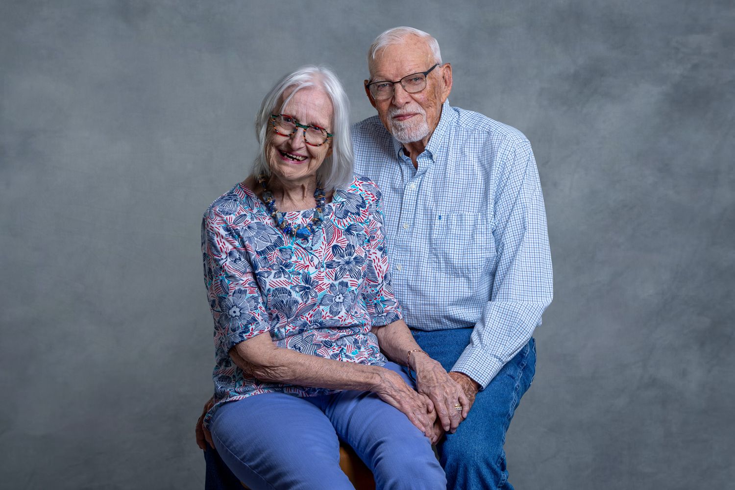 Elderly couple smiling and holding hands in a professional studio portrait with gray background.