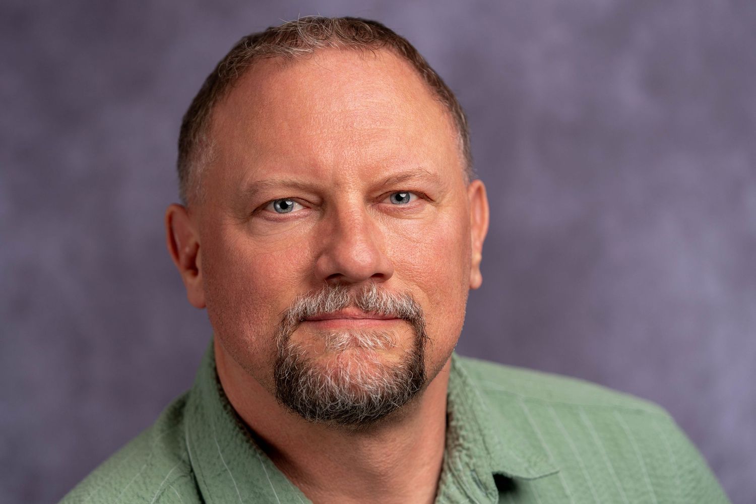 Close-up professional headshot of a bearded man in a green shirt against a gray backdrop.