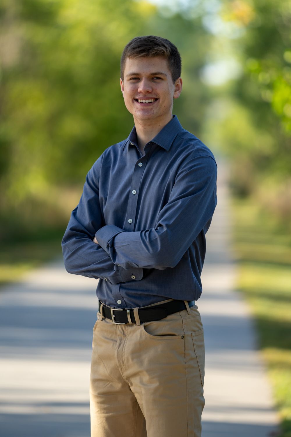 Smiling young man in blue button-up shirt standing with arms crossed on a tree-lined pathway outdoors.