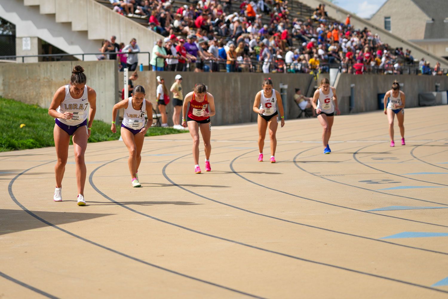 Female runners competing on an outdoor track in a crowded stadium during a collegiate track and field meet.