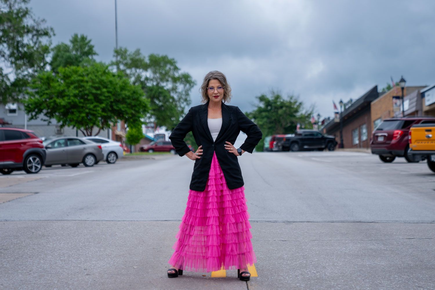 Woman in pink tulle skirt and black blazer stands confidently on street with hands on hips.