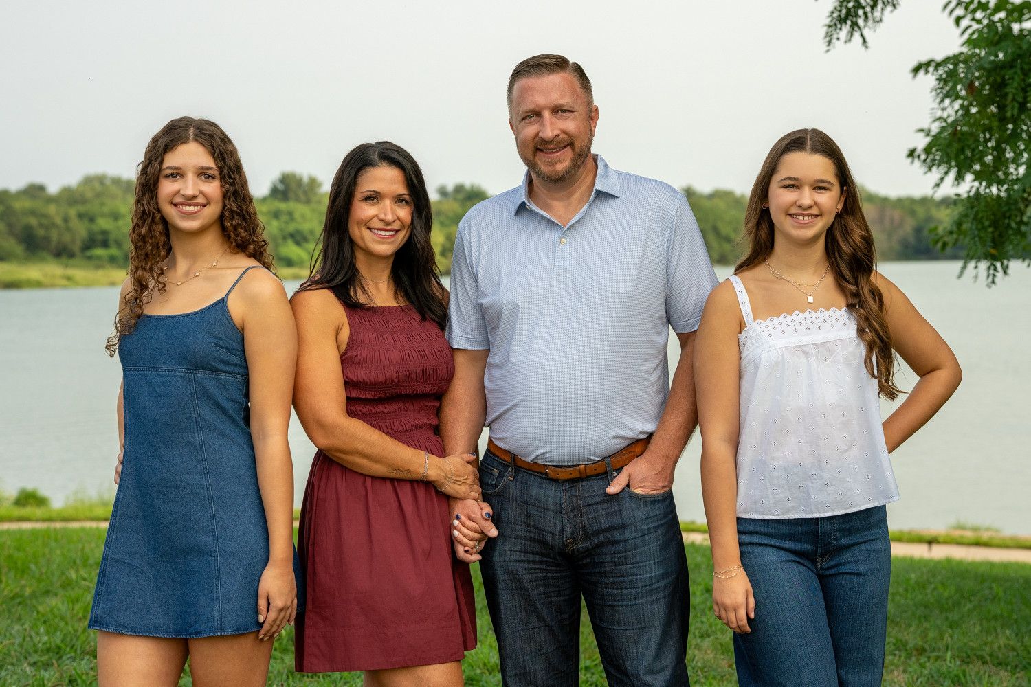 Family of four smiling outdoors near a lake, two teenage girls flanking their parents on a sunny day.